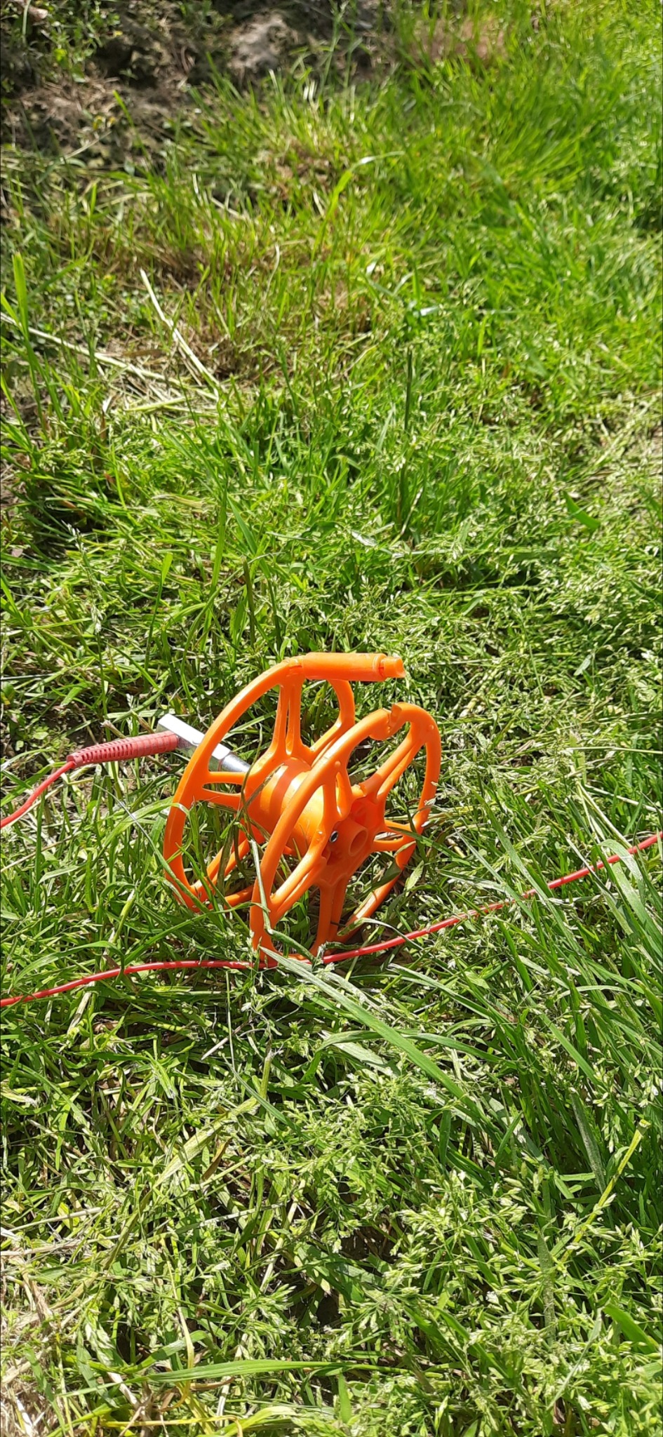 Pomarańczowy bęben z nawiniętym czerwonym przewodem, wbity w zieloną trawę, prawdopodobnie do uziemienia.
