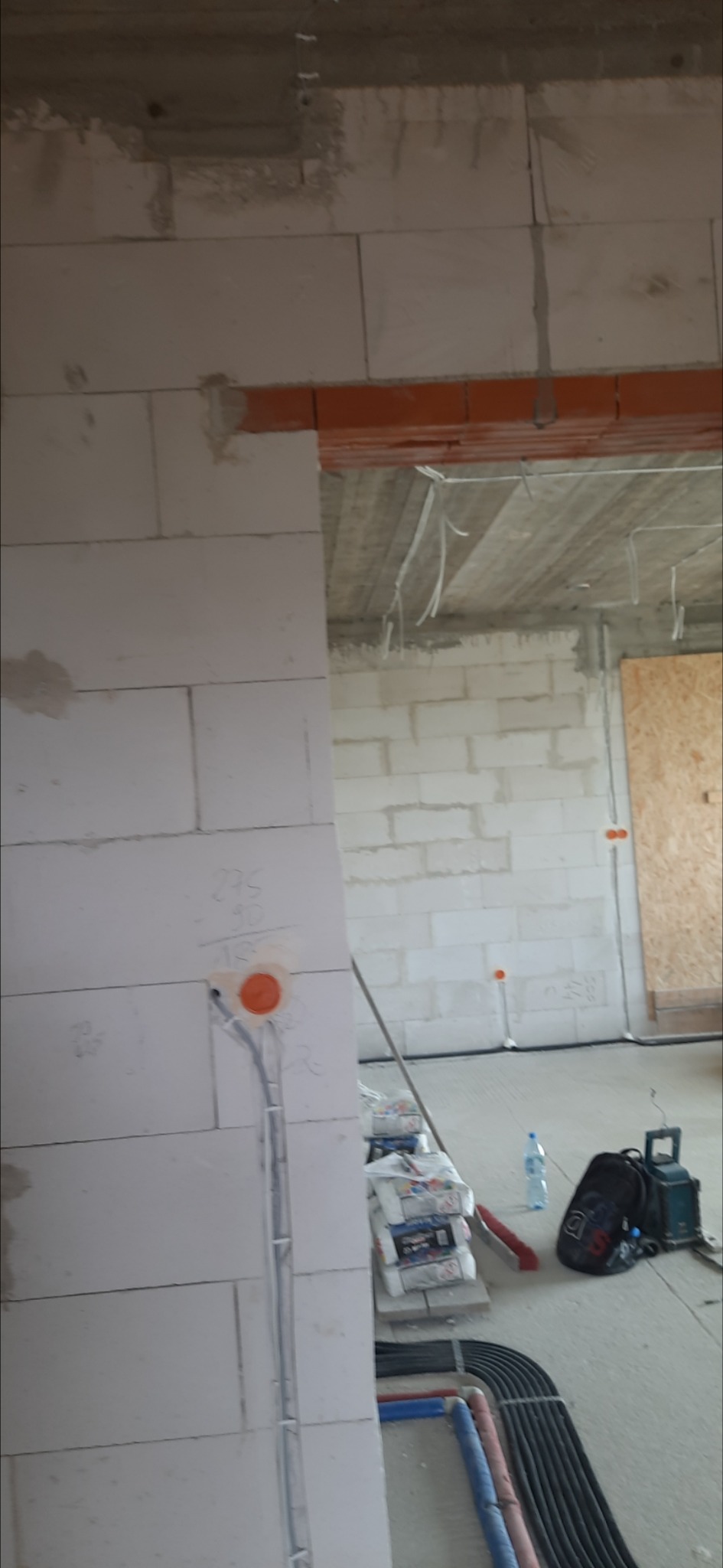 Wnętrze w trakcie prac instalacyjnych: widoczne przewody elektryczne w ścianie z betonu komórkowego, puszka elektryczna z pomarańczowym deklem, wiązka kabli w peszlach na podłodze, worki z zaprawą,...