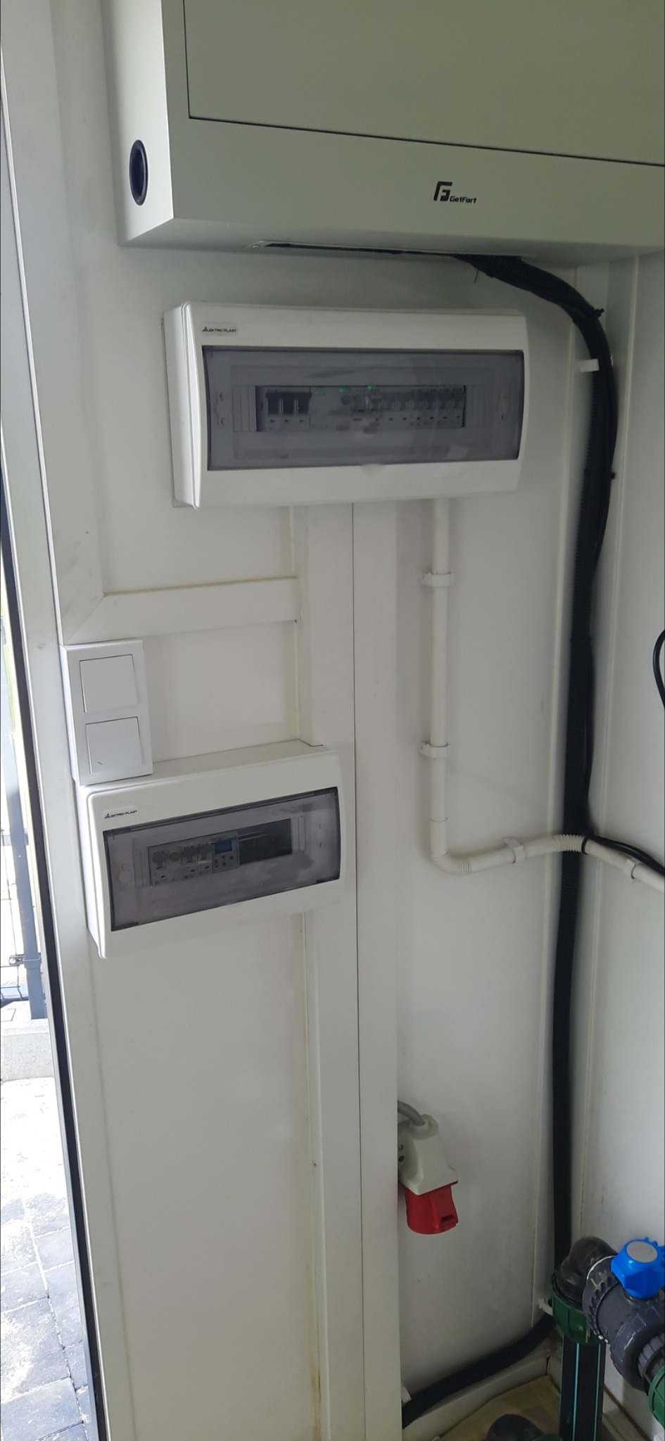 Dwa rozdzielnice elektryczne z bezpiecznikami zamontowane na białej ścianie, widoczne przewody w peszlu i włączniki światła, złącze siłowe w tle, oraz elementy instalacji wodnej na dole kadru.