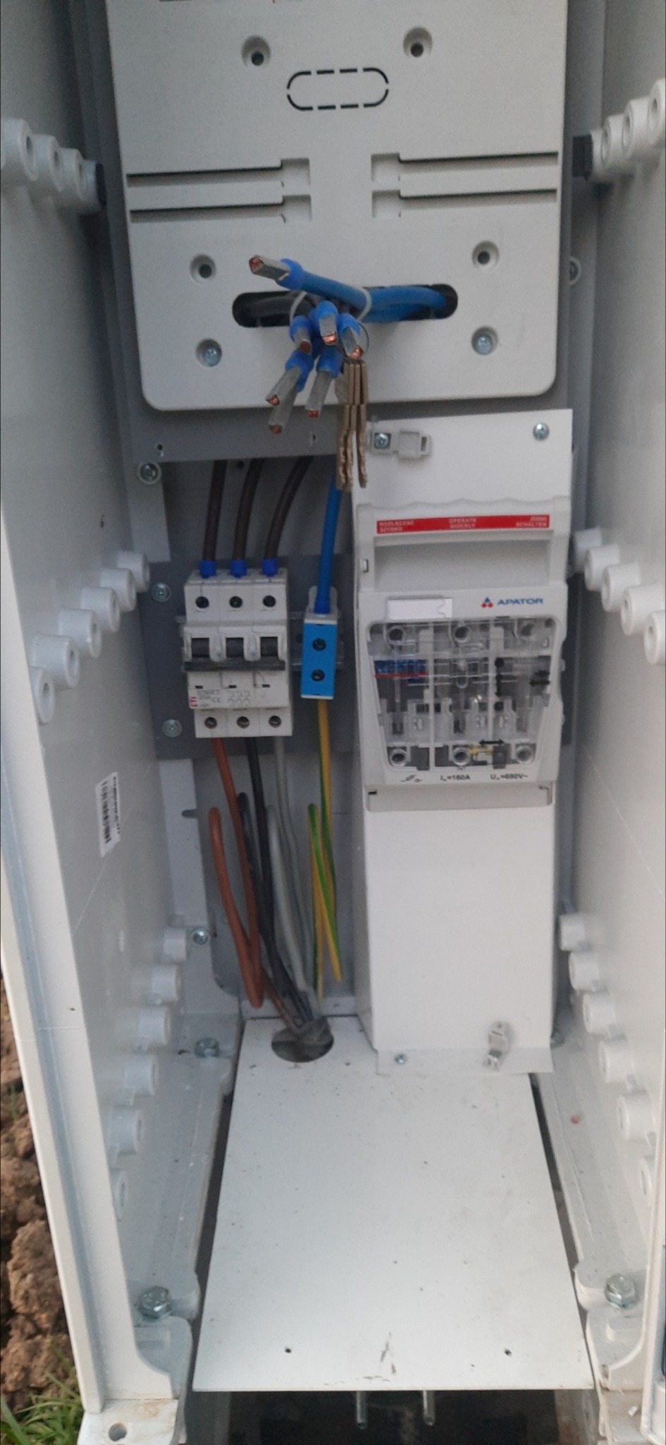 Wnętrze skrzynki elektrycznej z zamontowanym licznikiem Apator, bezpiecznikami i przewodami o różnych kolorach, w tym niebieskie z tulejkami, brązowe, czarne i żółto-zielone.