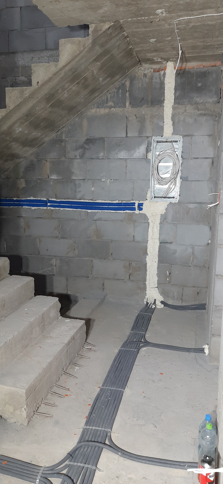 Instalacja elektryczna w surowym stanie: kable w peszlach prowadzone wzdłuż ściany z bloczków betonowych i pod schodami, puszka elektryczna zalana pianką montażową, widoczne fragmenty instalacji...
