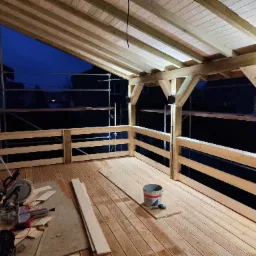 Zadaszony taras w trakcie budowy, widoczne drewniane elementy konstrukcyjne, deski podłogowe, narzędzia i materiały budowlane, wieczorne oświetlenie.