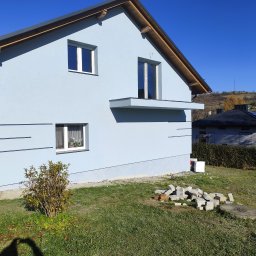 Krzysztof pierzynski - Jasnoniebieski dom jednorodzinny z białymi oknami i szarą dachówką w słoneczny dzień. Widoczne fragmenty ogrodu i sąsiednich budynków.