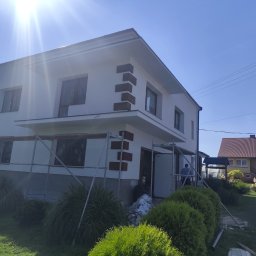 Krzysztof pierzynski - Biały dom z dekoracyjnymi, brązowymi elementami elewacji, częściowo zasłonięty rusztowaniem. Widoczne krzewy i sąsiedni dom w tle, jasne, słoneczne oświetlenie.