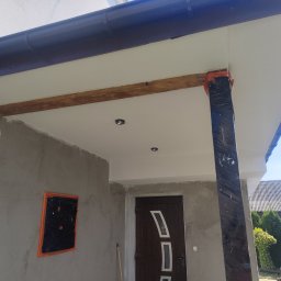 Krzysztof pierzynski - Zadaszenie wejścia do domu w trakcie prac wykończeniowych: surowy tynk, zabezpieczona folią kolumna, drewniana belka. Widoczne elementy elewacji i dachu.