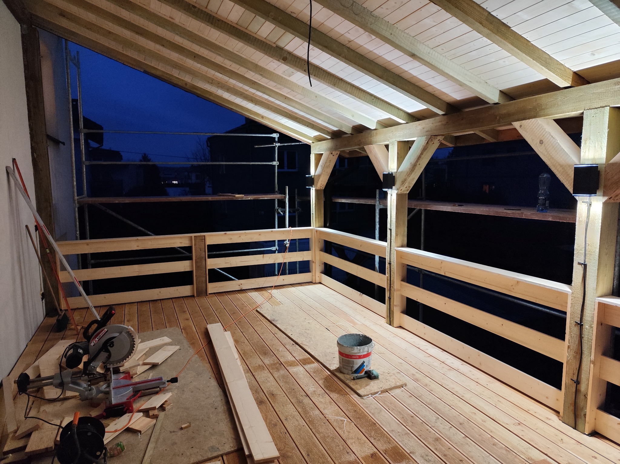 Zadaszony taras w trakcie budowy, widoczne drewniane elementy konstrukcyjne, deski podłogowe, narzędzia i materiały budowlane, wieczorne oświetlenie.