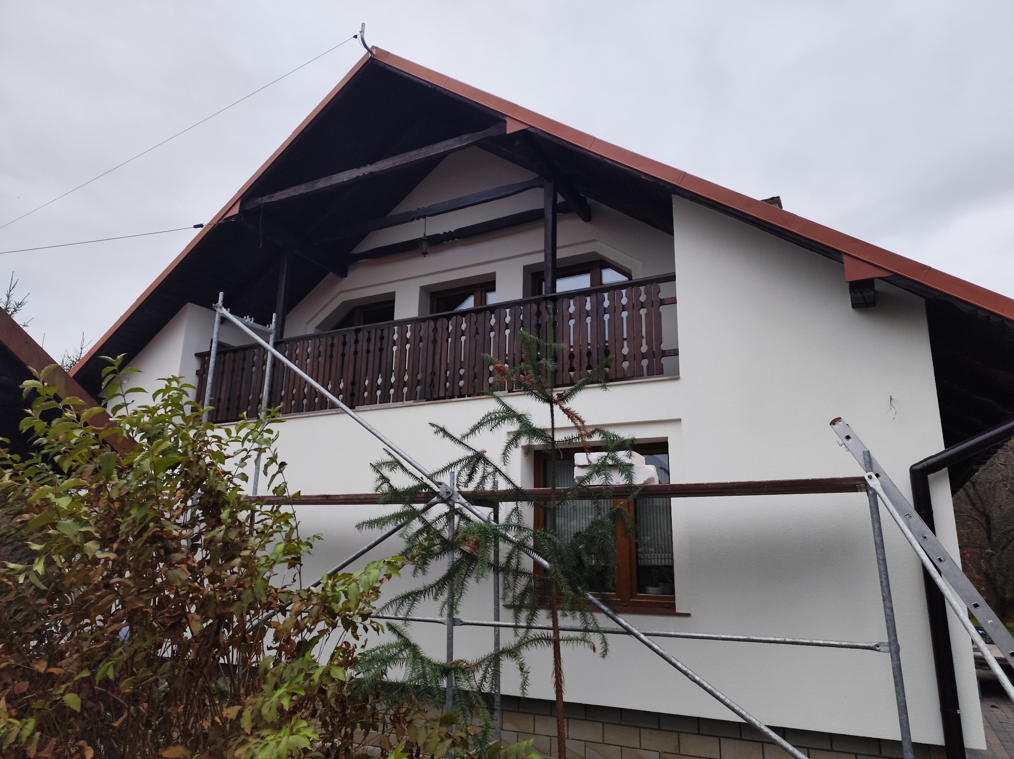 Dom z białym otynkowaniem, brązowym dachem i drewnianą balustradą balkonu, częściowo zasłonięty rusztowaniem i roślinnością, po wykonaniu elewacji.