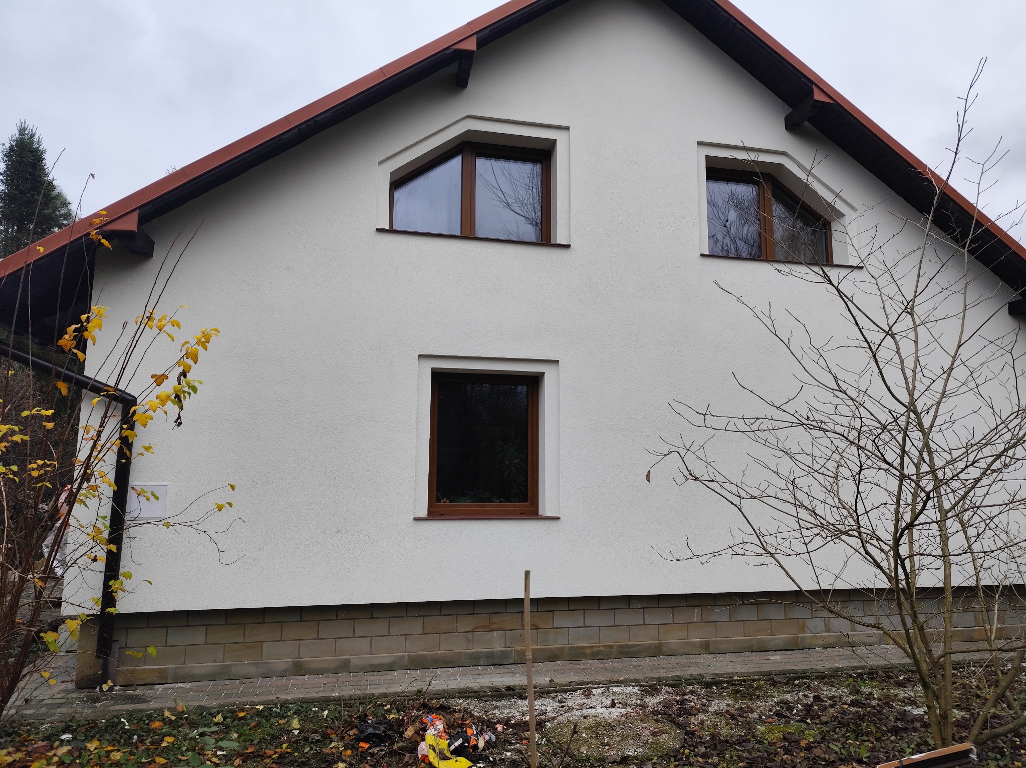 Biała elewacja domu jednorodzinnego z brązowymi oknami i dachem, widoczne jesienne drzewa i resztki liści na ziemi.