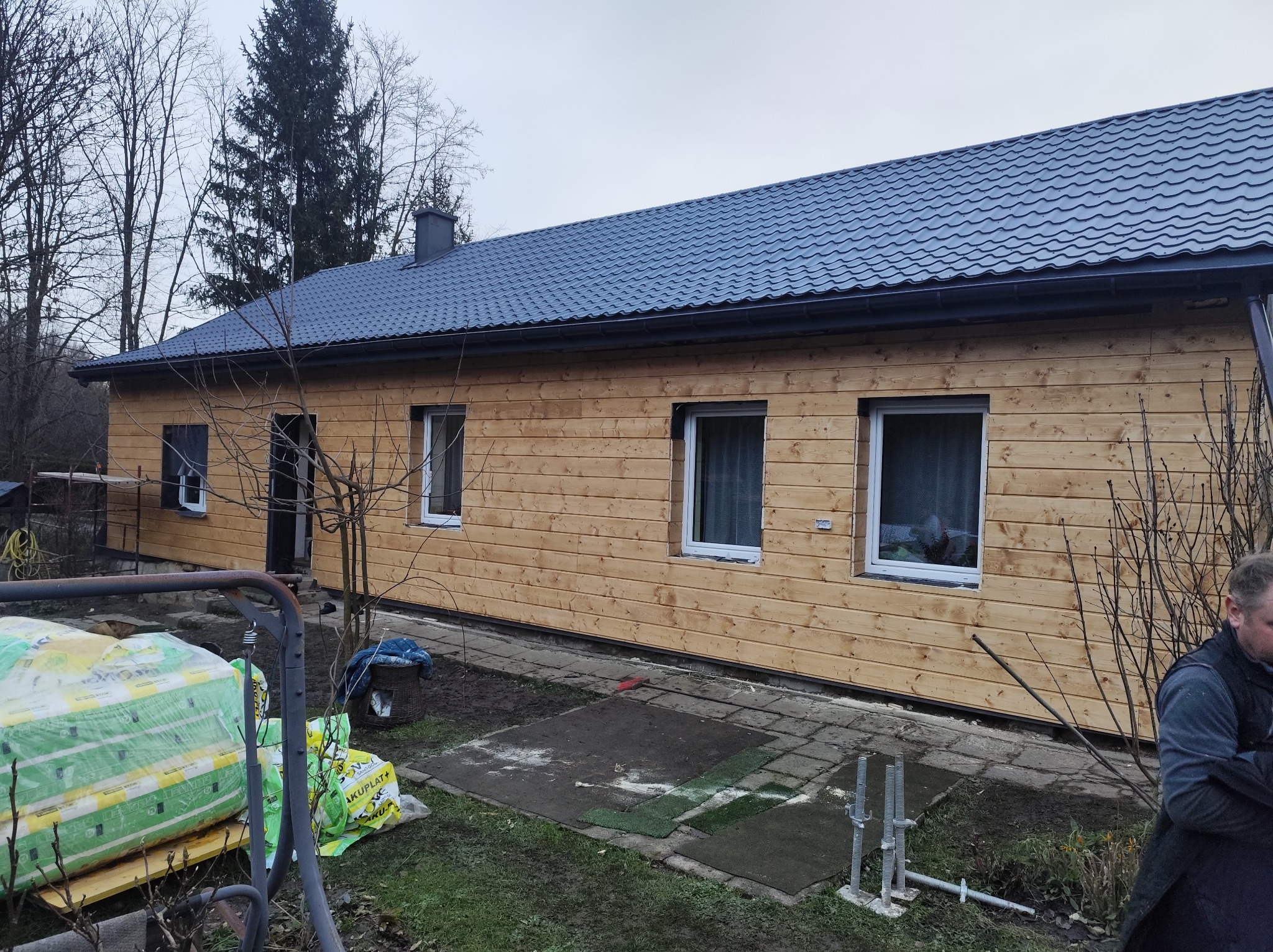 Dom z drewnianą elewacją i nowym, grafitowym dachem. Na pierwszym planie materiały budowlane i fragment osoby. Widoczne okna i komin. Pochmurne niebo w tle.