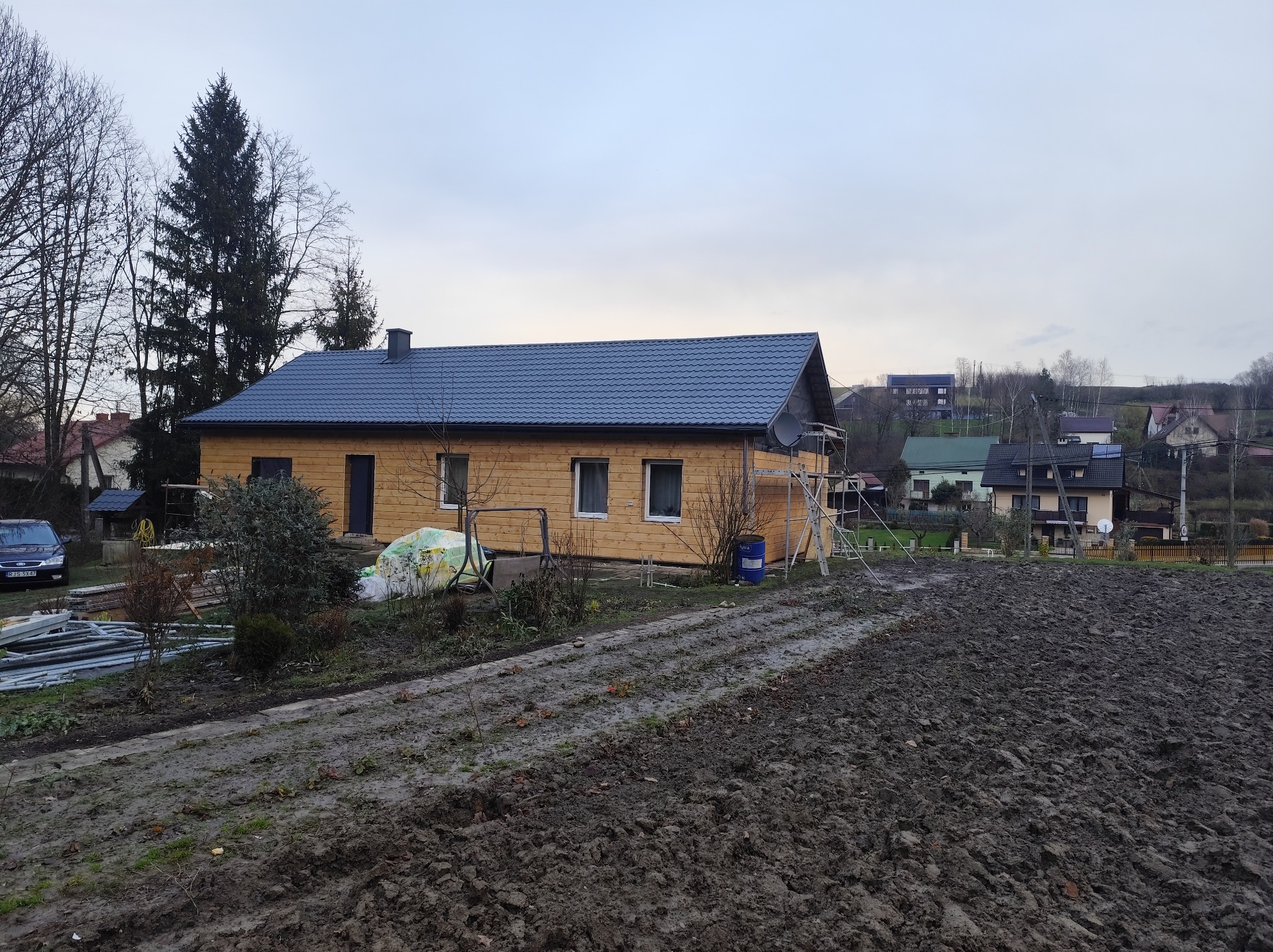 Drewniany dom z ciemnym dachem po remoncie elewacji, widoczny z perspektywy ogrodu. Na pierwszym planie świeżo przekopana ziemia, w tle inne zabudowania.