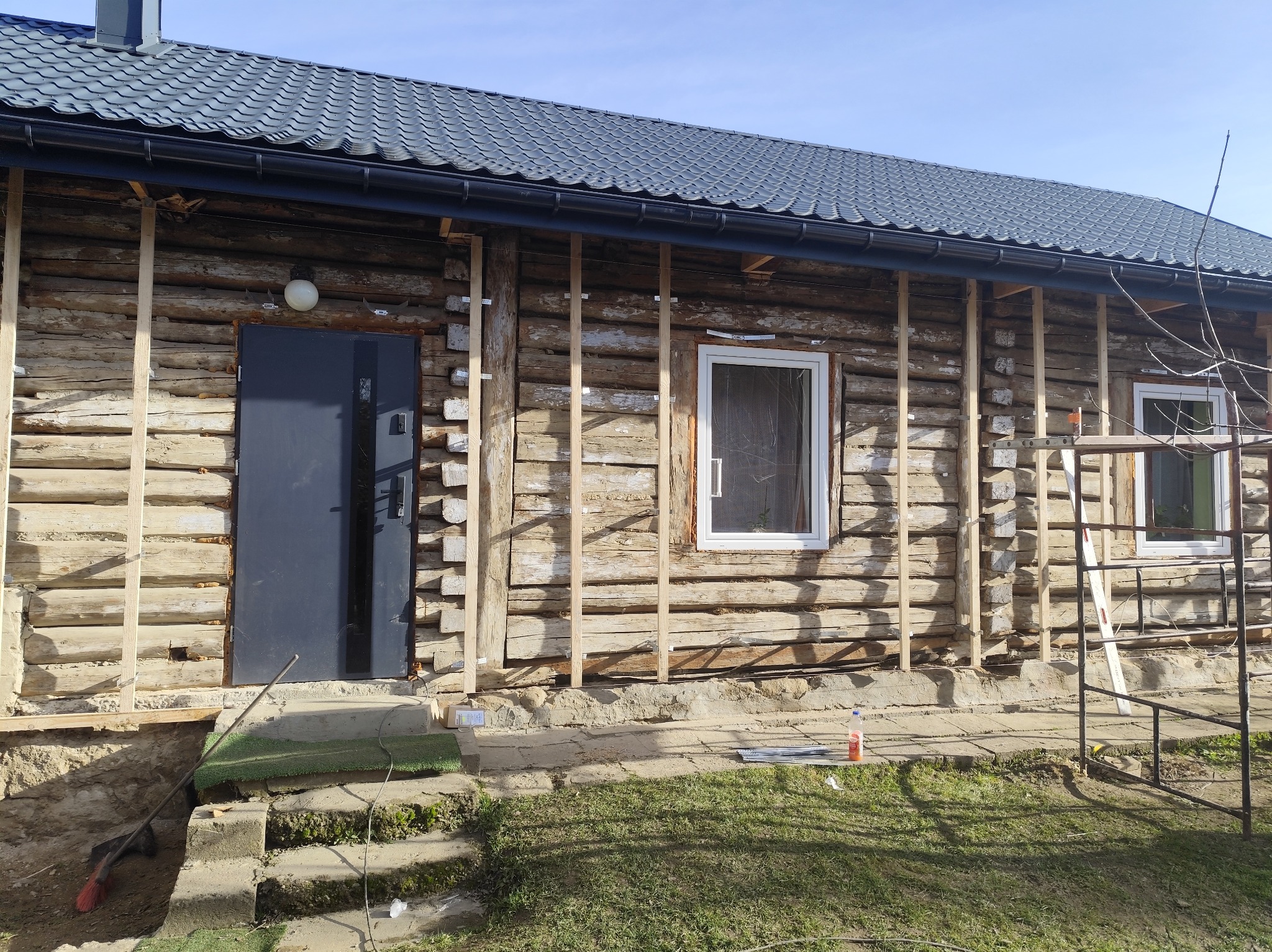 Dom z bali w trakcie przygotowania do ocieplenia. Widoczne drewniane listwy montażowe, okna z białymi ramami i metalowe rusztowanie. Dach pokryty blachodachówką.