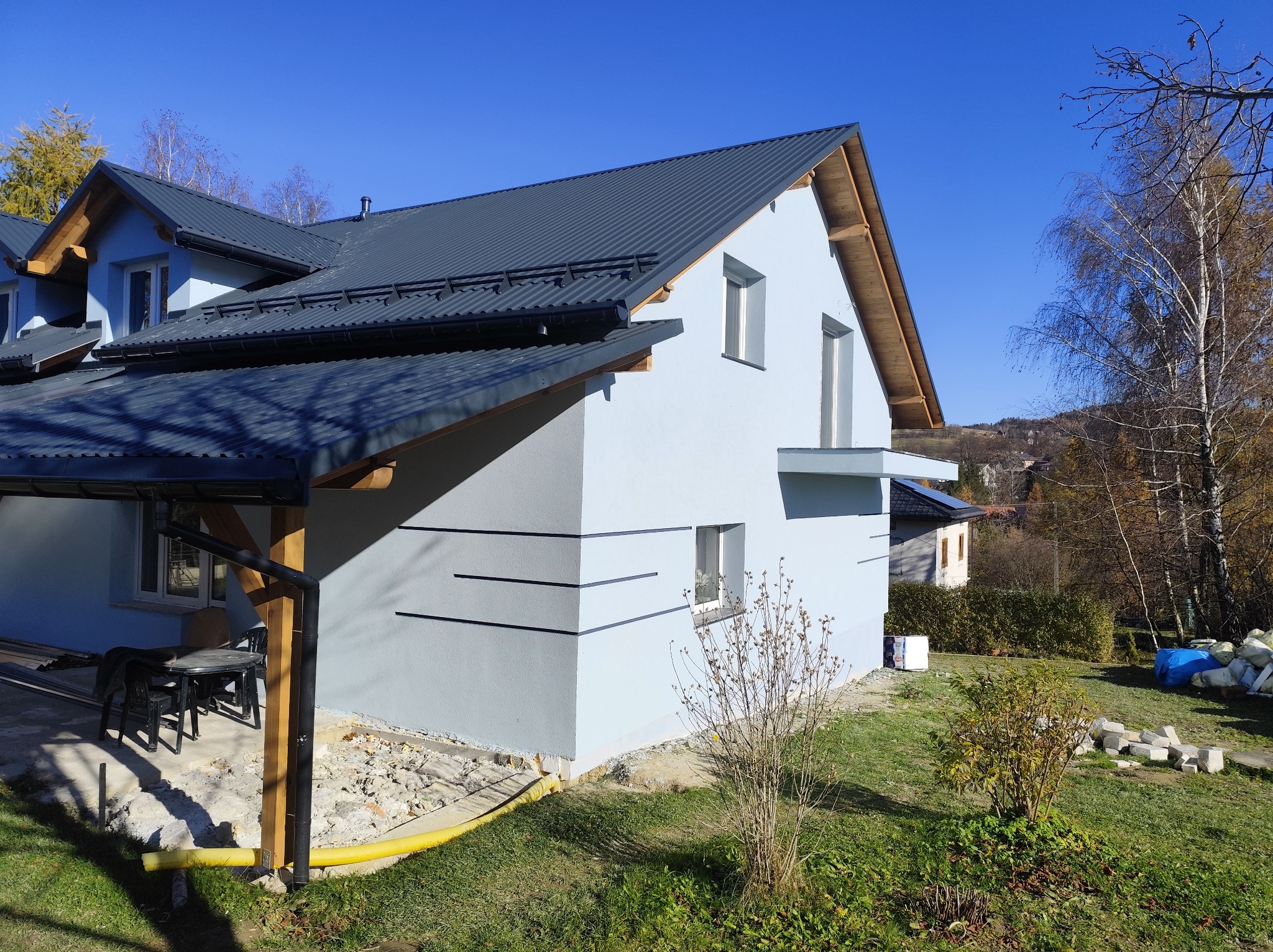 Jasny, otynkowany dom z ciemnym dachem blachodachówką, widoczny z boku, z fragmentem tarasu i ogrodu. Elementy dekoracyjne na elewacji. Częściowo wykończony teren.