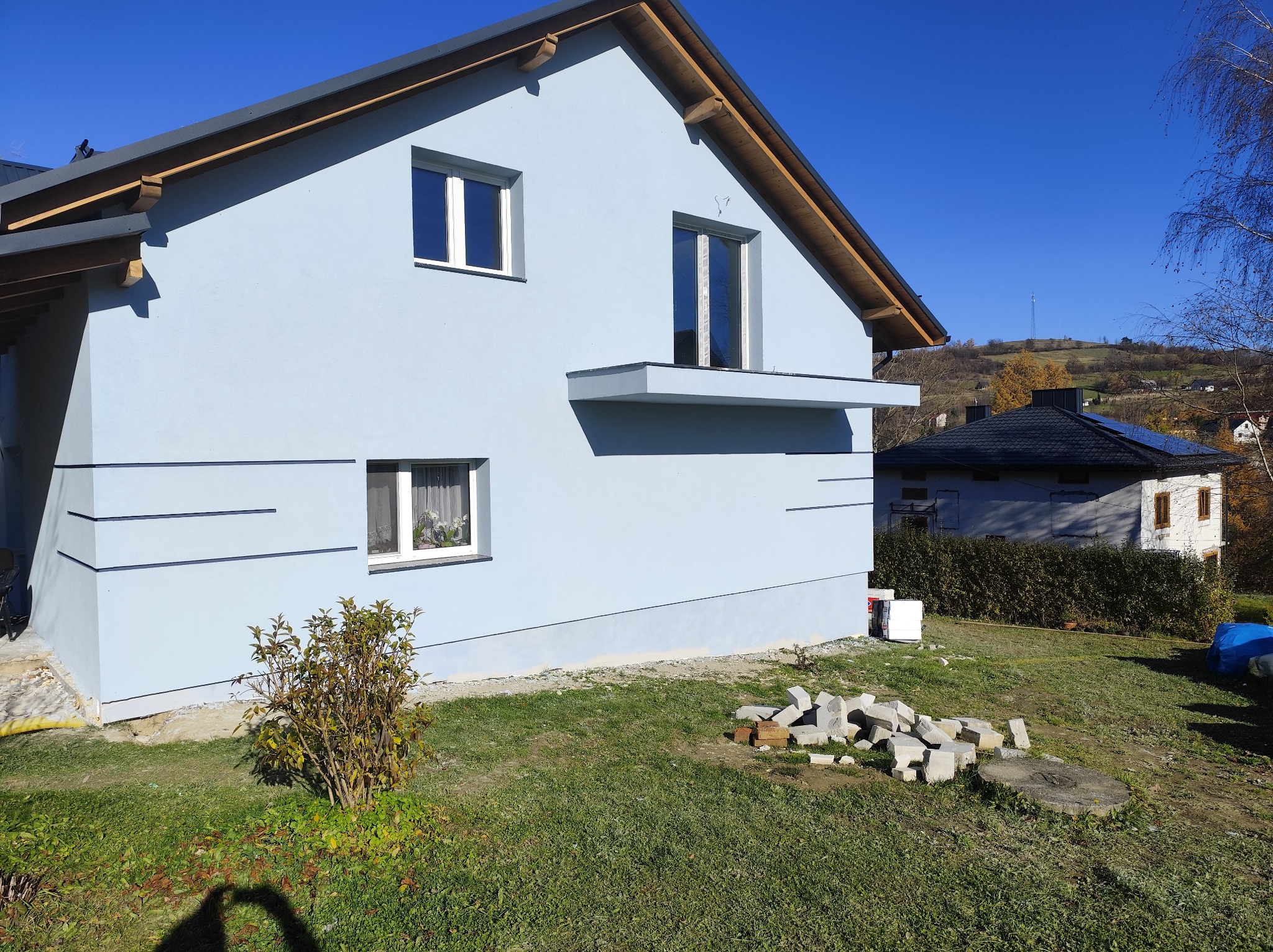 Jasnoniebieski dom jednorodzinny z białymi oknami i szarą dachówką w słoneczny dzień. Widoczne fragmenty ogrodu i sąsiednich budynków.