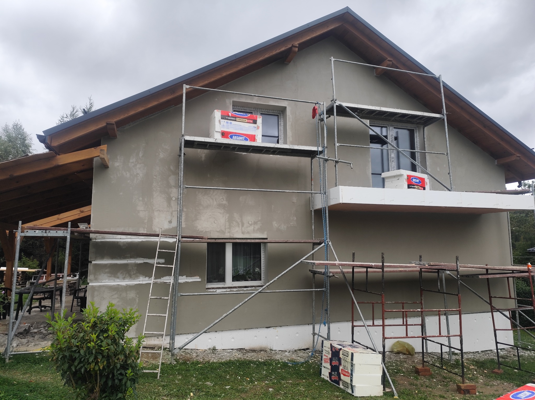 Dom w trakcie ocieplania: styropian, rusztowania, szara elewacja. Widoczne pakiety styropianu Arsanit. Częściowo otynkowana ściana. Ujęcie z poziomu gruntu.