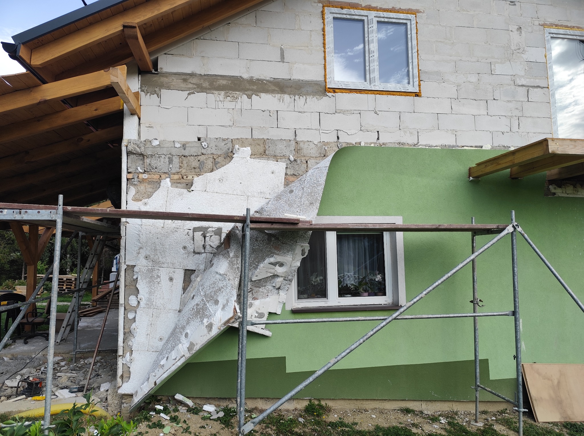 Fragment ocieplanej elewacji domu z widocznymi rusztowaniami, styropianem i fragmentem pomalowanej ściany na zielono. Widoczne okna i drewniana konstrukcja dachu.