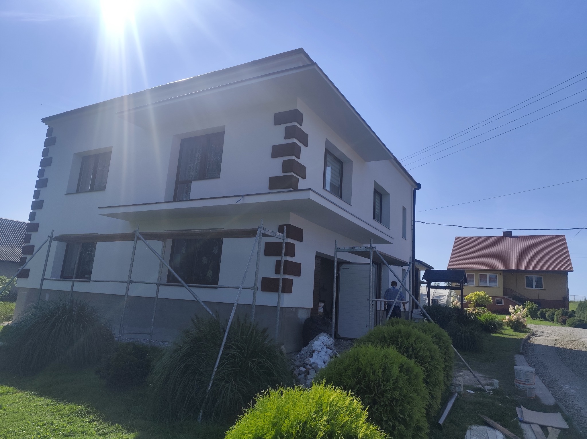 Biały dom z dekoracyjnymi, brązowymi elementami elewacji, częściowo zasłonięty rusztowaniem. Widoczne krzewy i sąsiedni budynek w tle, jasne, słoneczne oświetlenie.