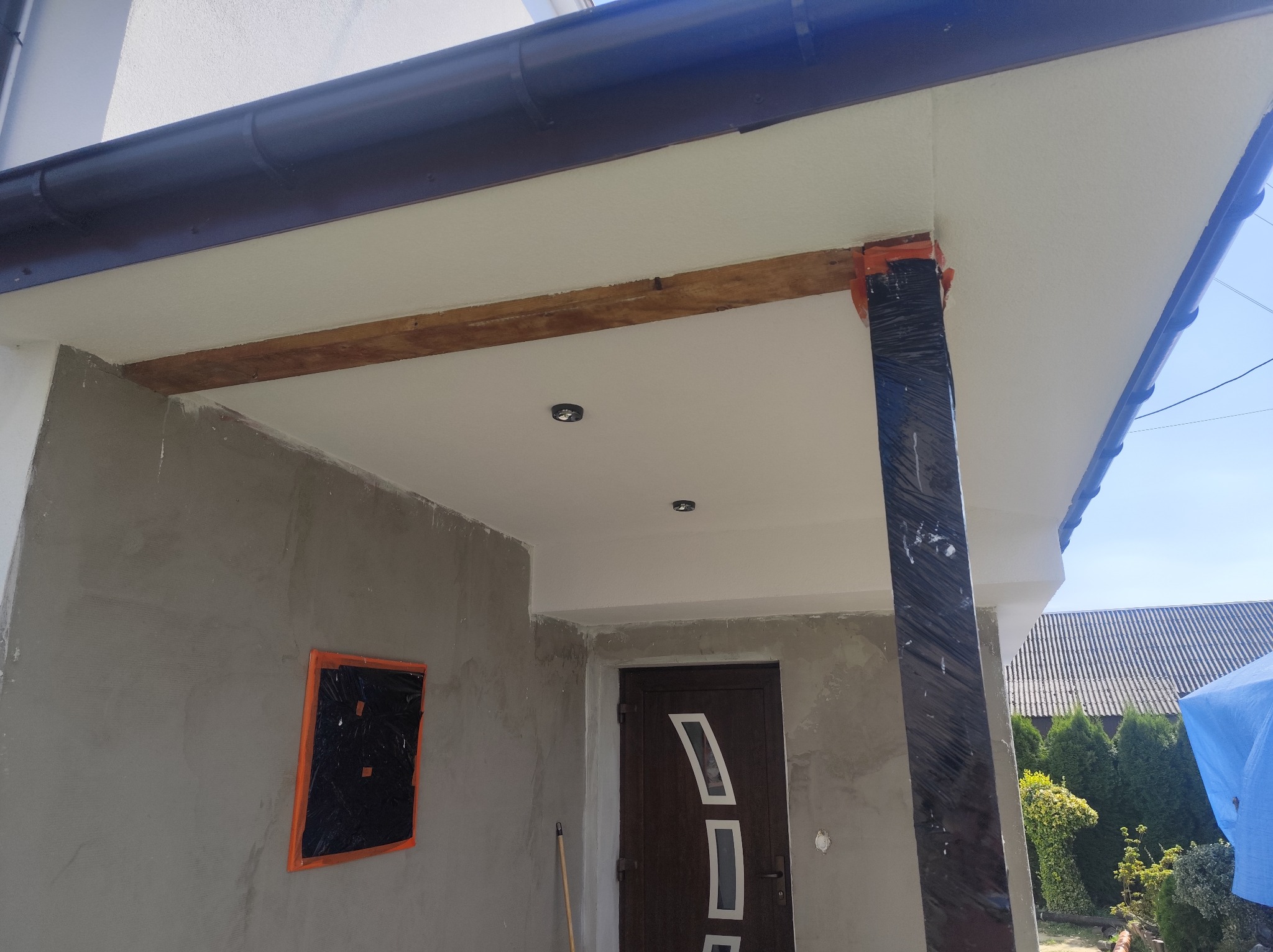 Zadaszenie wejścia do domu w trakcie prac wykończeniowych: surowy tynk, zabezpieczona folią kolumna, drewniana belka. Widoczne elementy elewacji i dachu.
