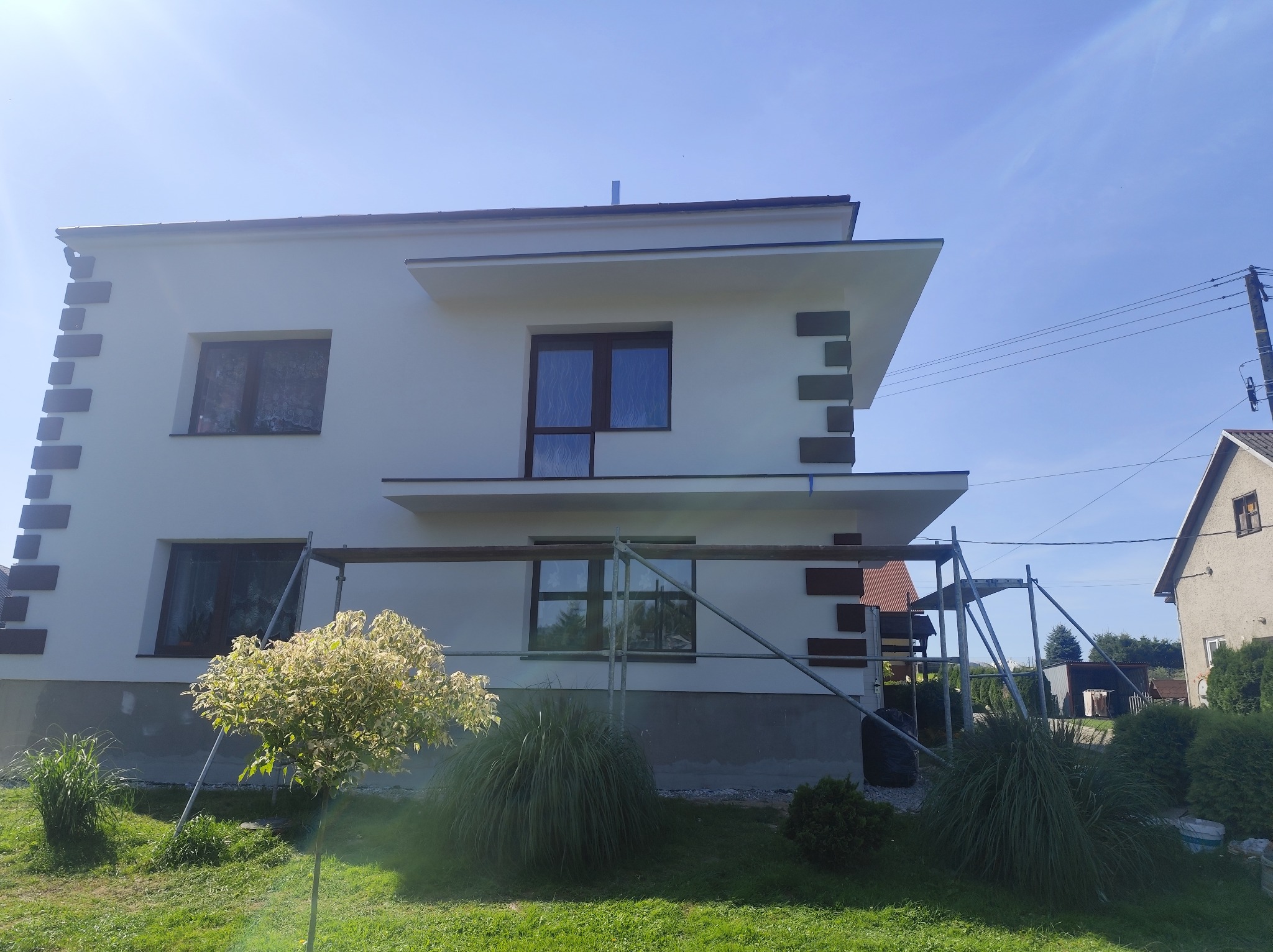 Dwukondygnacyjny dom z białą elewacją i dekoracyjnymi, ciemnymi elementami. Widoczne rusztowania sugerują prace wykończeniowe.