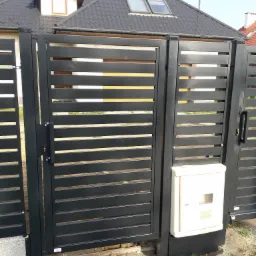 Nowoczesna, czarna brama wjazdowa z poziomymi szczeblami, zamontowana przy domu z grafitowym dachem i beżową skrzynką na gaz.