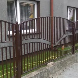 Brązowe ogrodzenie z metalowymi pionowymi prętami i dekoracyjnymi, falistymi elementami, widoczne okna domu w tle.