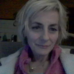 Portret uśmiechniętej kobiety z krótkimi blond włosami, ubrana w różowy szal i jasny sweter, z półką z dekoracjami w tle.