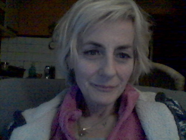 Portret uśmiechniętej kobiety z krótkimi blond włosami, ubrana w różowy szal i jasny sweter, z półką z dekoracjami w tle.