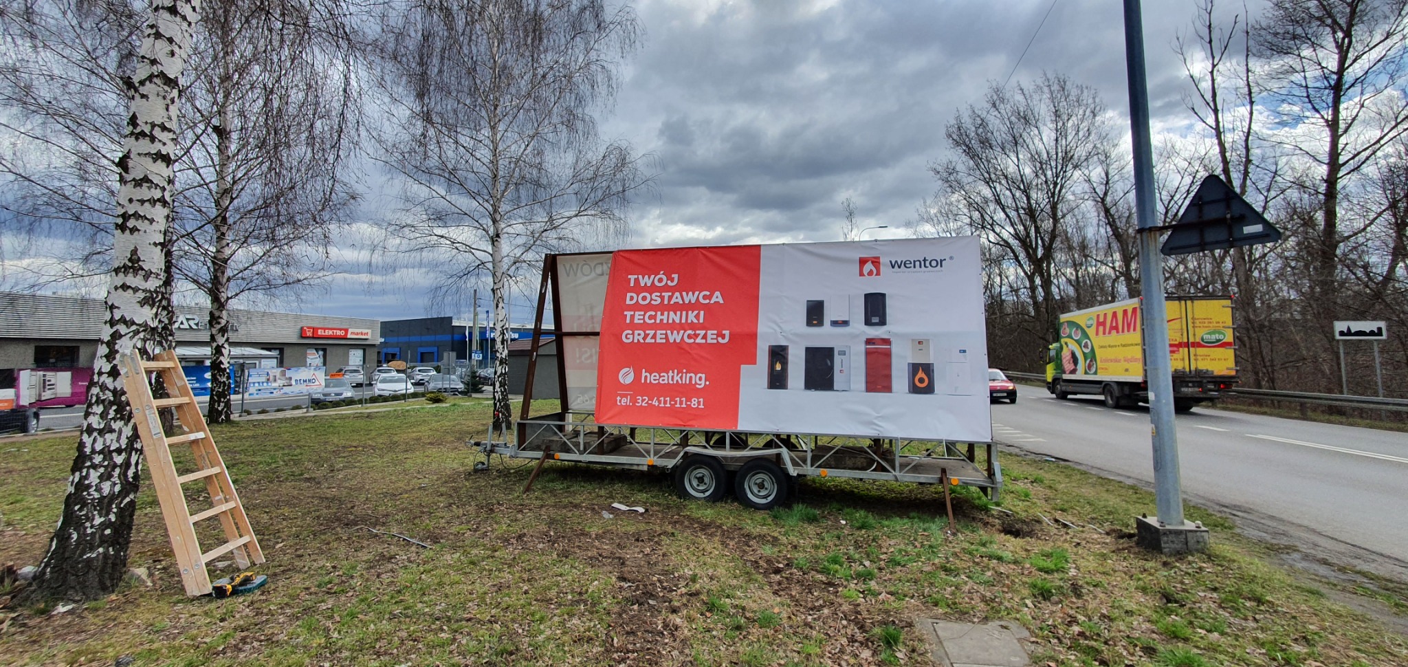 Przyczepa reklamowa z banerem prezentującym urządzenia grzewcze Wentor i Heatking, ustawiona obok drogi, z drabiną opartą o brzozę w tle.