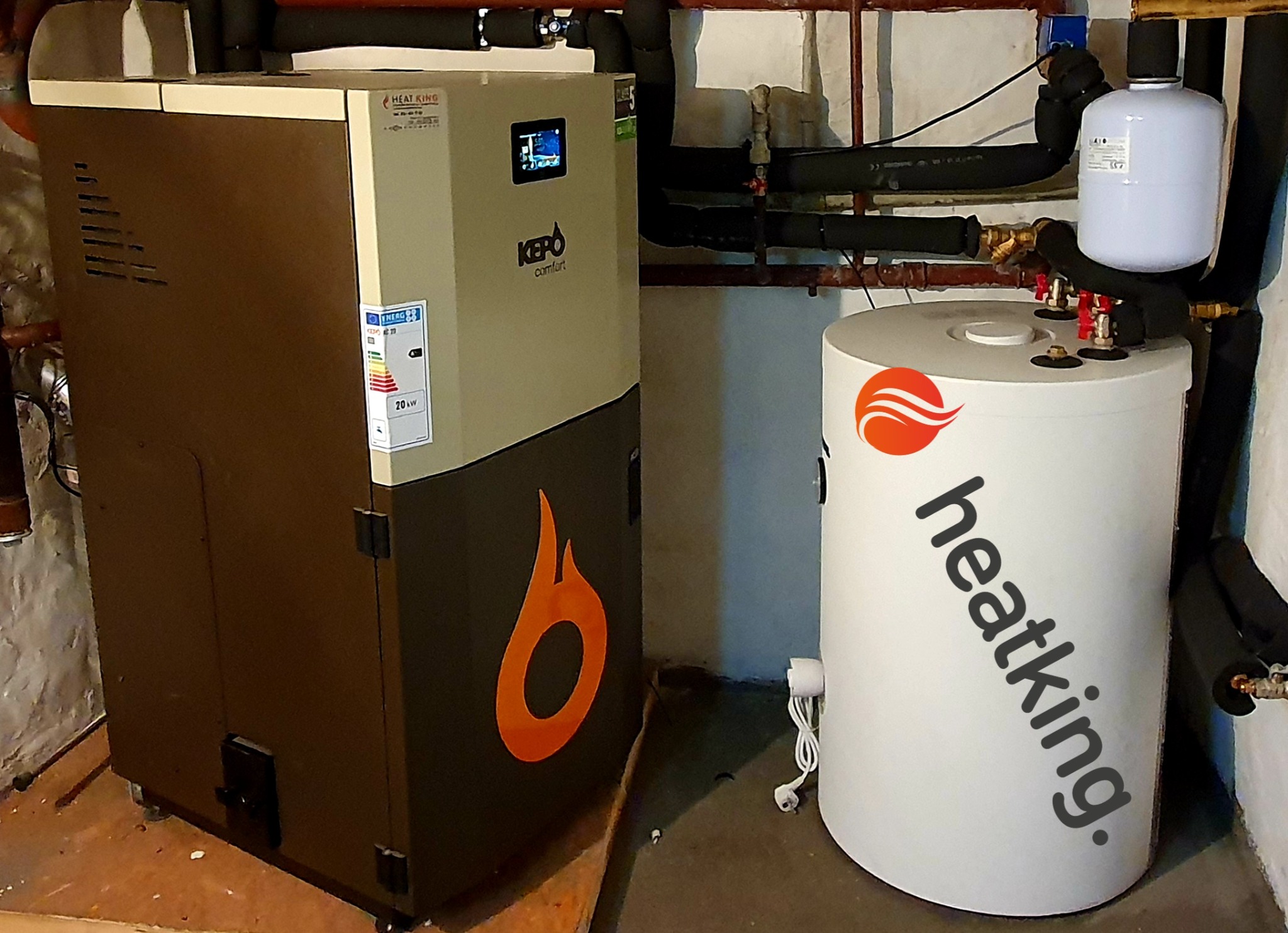 Kocioł na paliwo stałe Kepo Comfort z wyświetlaczem i etykietą energetyczną, obok biały zbiornik Heatking z instalacją hydrauliczną w piwnicy.