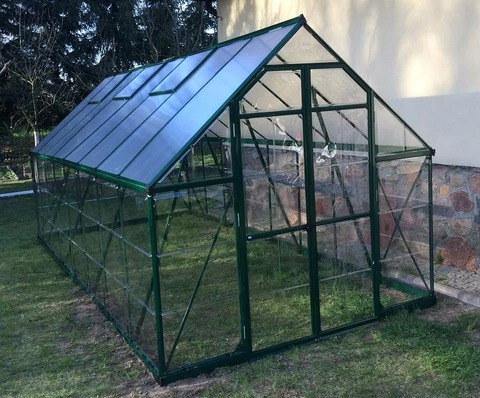 Szklarnia z zielonymi ramami na trawniku, z otwartymi oknami dachowymi, na tle ściany budynku z kamienną podmurówką.