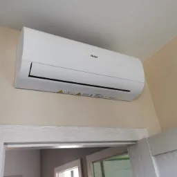 Biały klimatyzator Haier zamontowany na kremowej ścianie nad futryną drzwiową z szarego drewna.