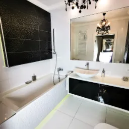 Nowoczesna łazienka w czarno-białej kolorystyce z wanną obudowaną białą, teksturowaną mozaiką, czarną ścianą z płytek o chropowatej fakturze, białą umywalką nablatową i czarnym oświetleniem w stylu...