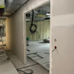 Wnętrze w trakcie prac wykończeniowych: widoczne płyty kartonowo-gipsowe na ścianach, elementy instalacji wentylacyjnej pod sufitem, oraz fragment podłogi z otworami rewizyjnymi i widoczną izolacją...