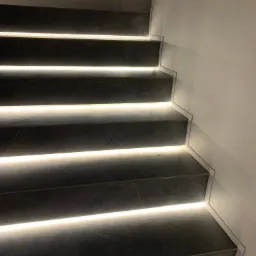 Schody wykończone ciemnym kamieniem z subtelnym podświetleniem LED na każdym stopniu, oświetlające białą ścianę obok.