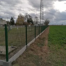 Nowo zamontowane, zielone ogrodzenie panelowe na betonowej podmurówce, oddzielające trawnik od pola uprawnego, z wieżą telekomunikacyjną w tle pod pochmurnym niebem.