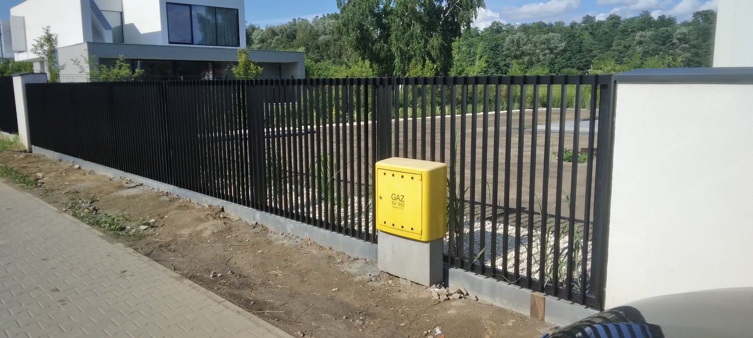 Czarny, pionowy płot metalowy z żółtą skrzynką gazową na betonowym fundamencie, na tle nowoczesnego domu i drzew