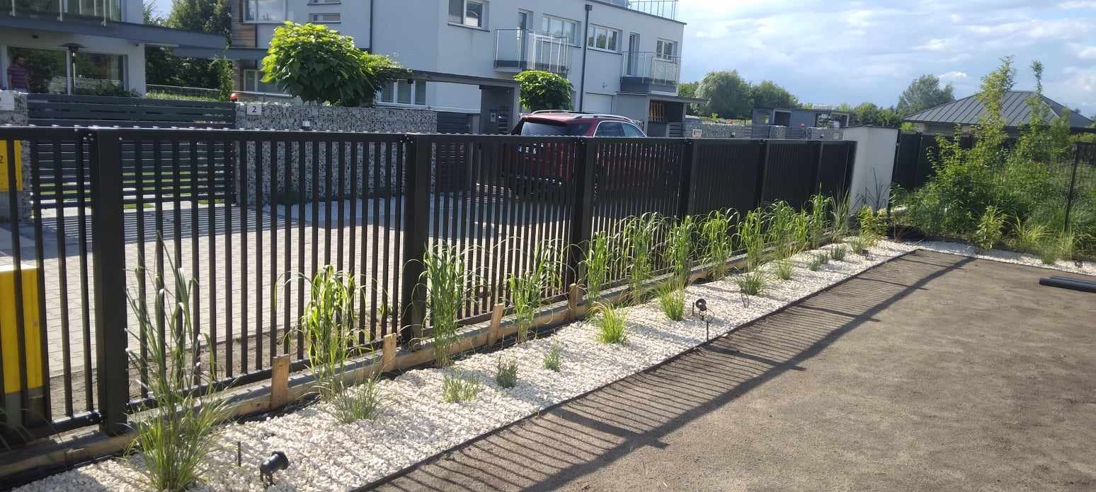 Nowoczesne ogrodzenie panelowe w kolorze czarnym, otoczone ozdobnymi trawami i białym żwirem, z widocznym fragmentem budynku w tle.