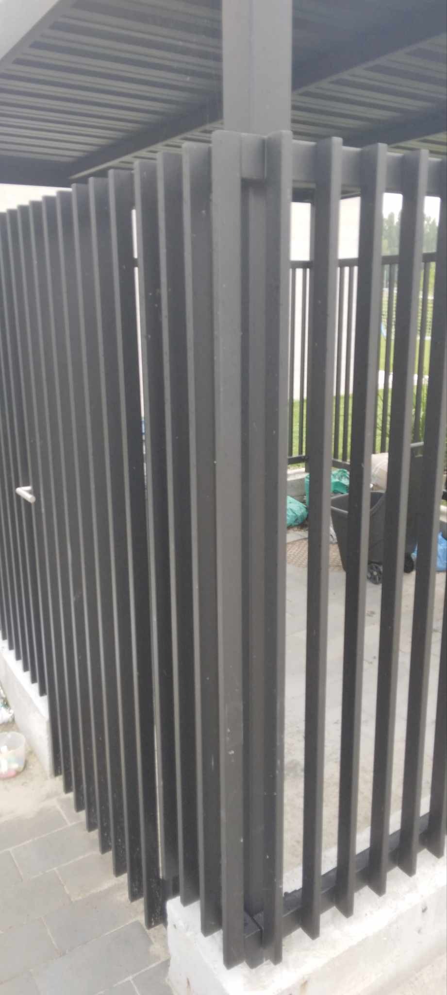 Nowoczesne, pionowe, metalowe ogrodzenie w kolorze grafitowym, z widoczną konstrukcją zadaszenia i elementami otoczenia.