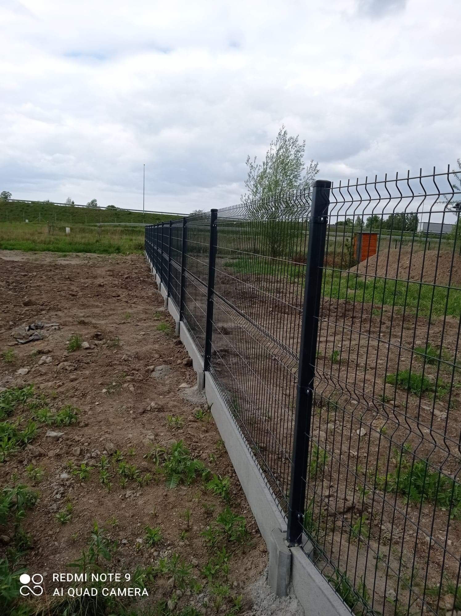 Długie, proste ogrodzenie z paneli zgrzewanych na betonowej podmurówce, widok z perspektywy ziemi w pochmurny dzień. W tle widoczne pole i drzewa.