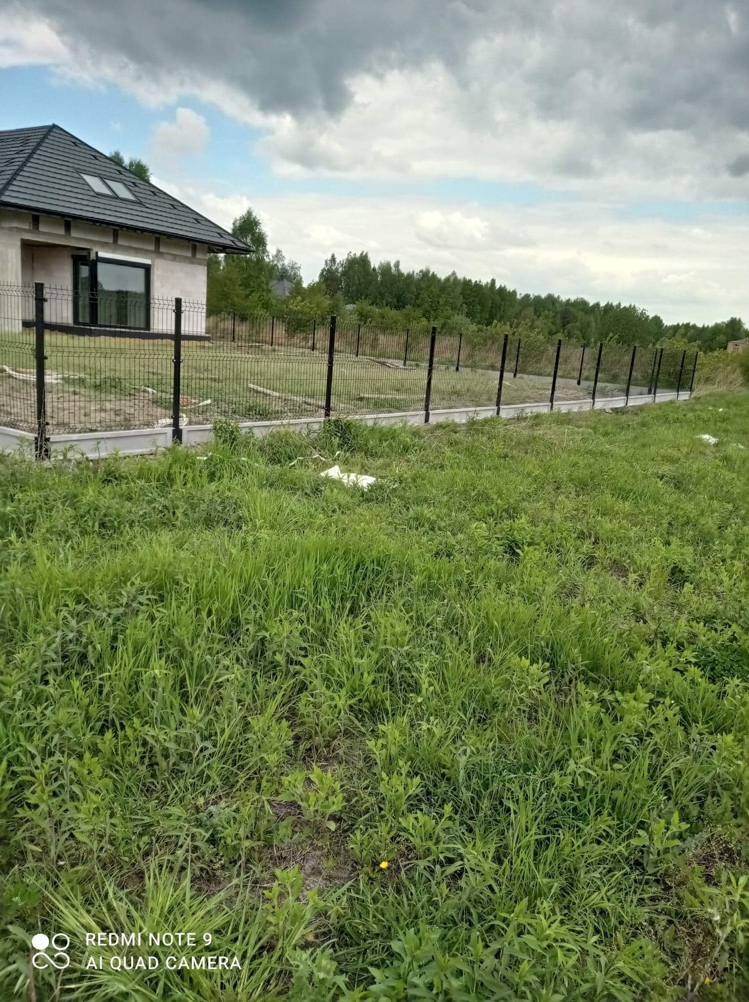 Nowoczesny dom z szarą dachówką otoczony czarnym ogrodzeniem panelowym na betonowej podmurówce, widok z zarośniętego trawnika w pochmurny dzień.