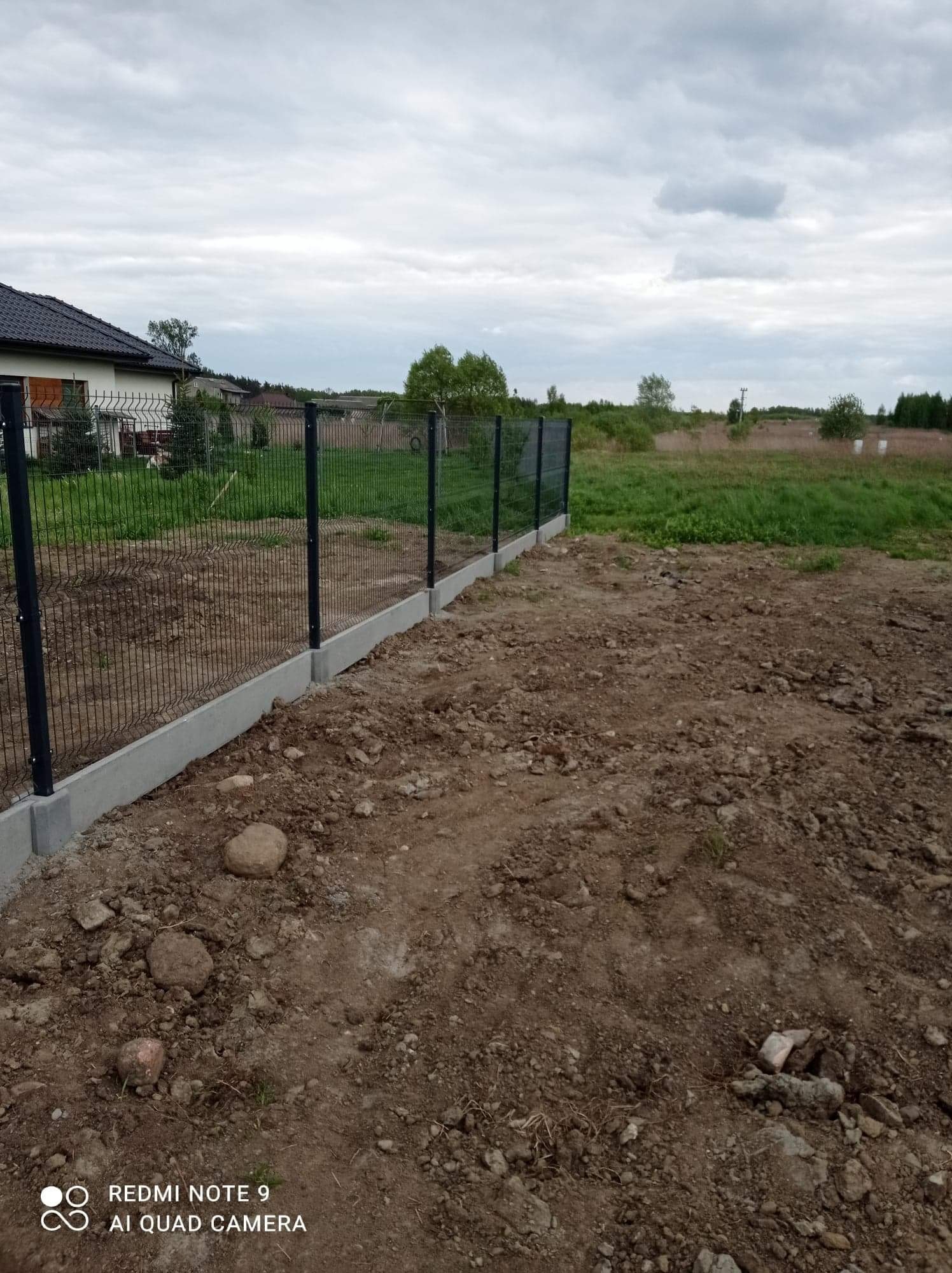 Nowo postawione ogrodzenie panelowe z podmurówką betonową na nierównym, przygotowywanym terenie działki, widok na dom i pole w pochmurny dzień.