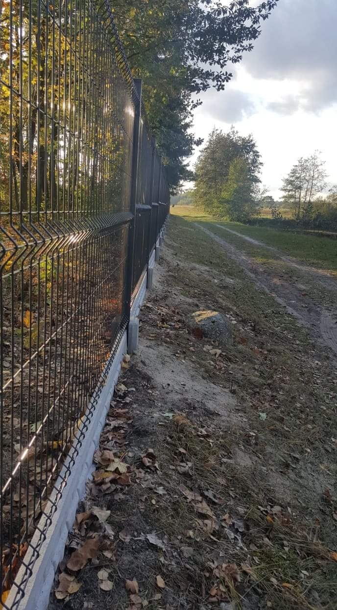 Długi, ciemny, metalowy płot z paneli na betonowej podmurówce, biegnący wzdłuż polnej drogi z drzewami w tle, widziany z perspektywy ziemi.