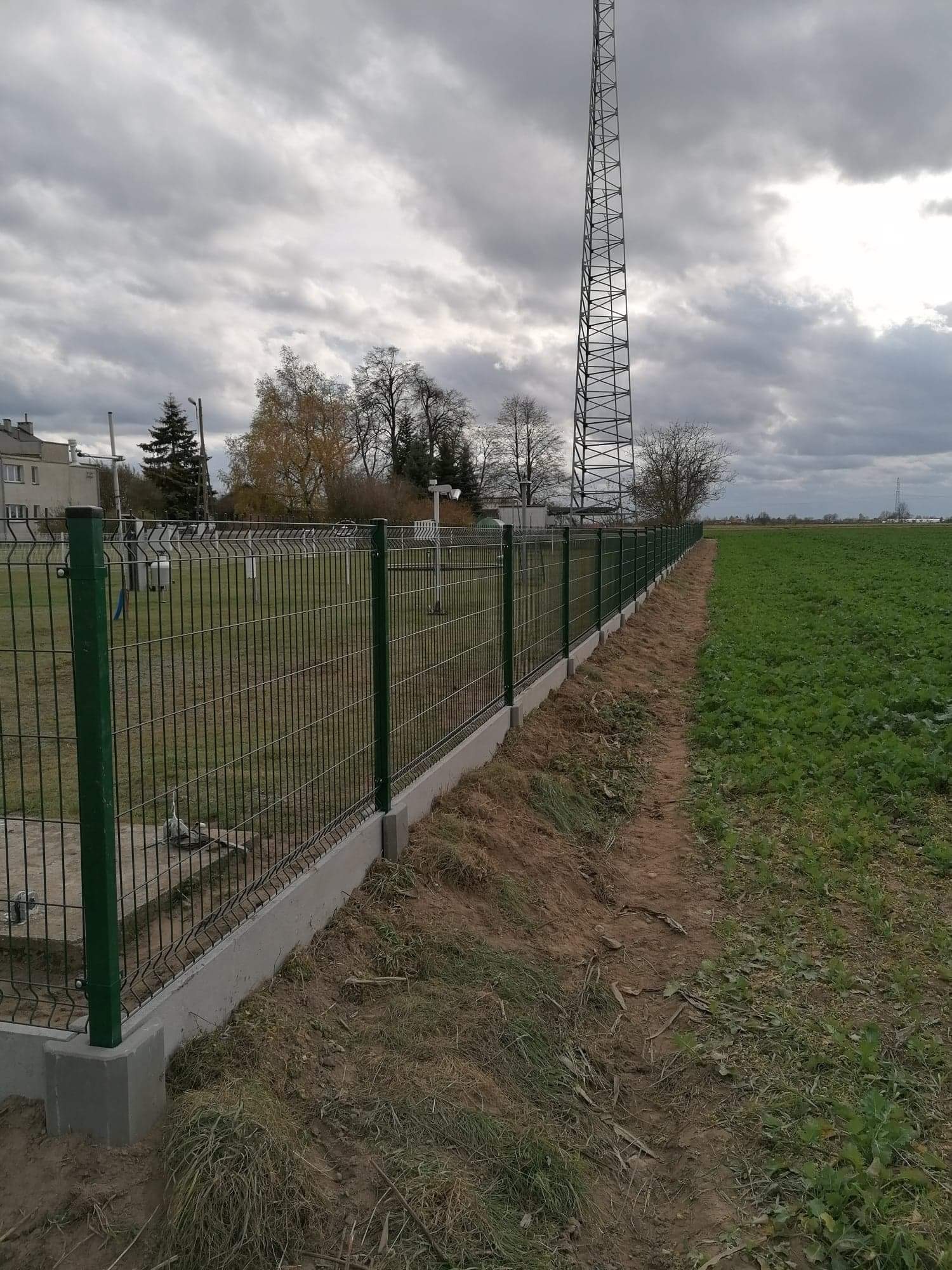 Nowo zamontowane, zielone ogrodzenie panelowe na betonowej podmurówce, oddzielające trawnik od pola uprawnego, z wieżą telekomunikacyjną w tle pod pochmurnym niebem.