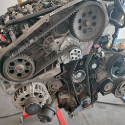 Mr Cars - Zdemontowany silnik samochodowy z widocznymi paskami rozrządu i kołami pasowymi, umieszczony na stojaku w warsztacie w Chrząstowicach. Widoczne ślady użytkowania i zabrudzenia na elementach.