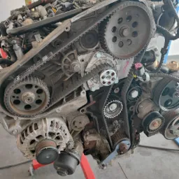 Zdemontowany silnik samochodowy z widocznymi paskami rozrządu i kołami pasowymi, umieszczony na stojaku w warsztacie w Chrząstowicach. Widoczne ślady użytkowania i zabrudzenia na elementach.