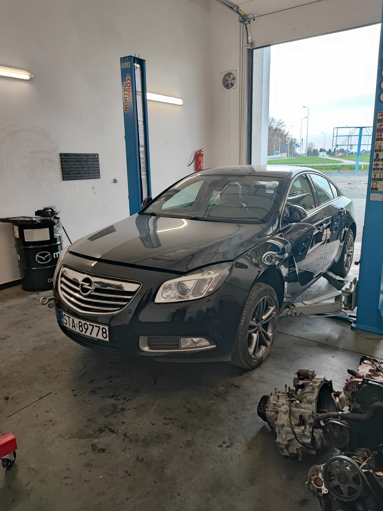 Czarny Opel Insignia na podnośniku w warsztacie, widoczny silnik na podłodze, beczka z olejem Mazda w tle, tablica rejestracyjna STA 89778