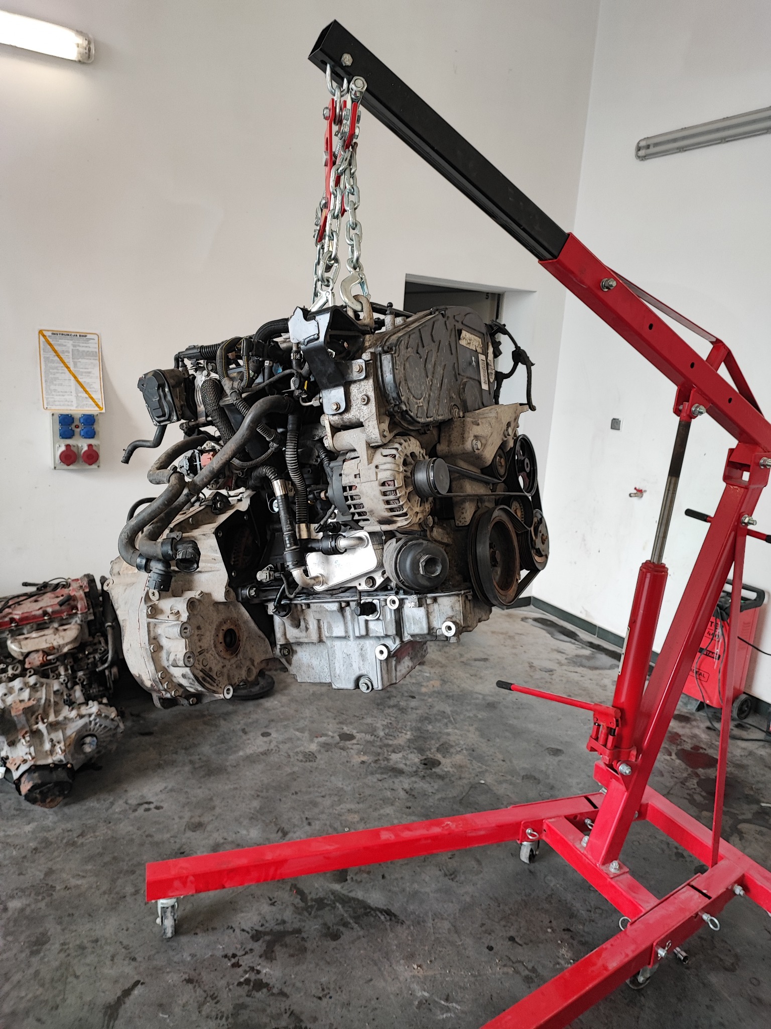 Silnik samochodowy zawieszony na łańcuchu z czerwonym podnośnikiem warsztatowym w tle; obok leżą inne zdemontowane silniki i skrzynie biegów; widoczne gniazda elektryczne na ścianie.