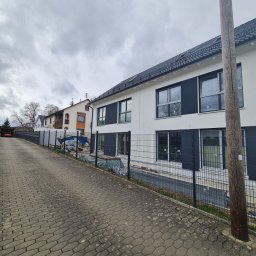 BauTech - Nowoczesny dom w Szczecinie z białą elewacją i grafitowymi akcentami, widoczny z boku posesji, z elementami ogrodzenia panelowego i brukowaną drogą dojazdową.