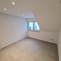 BauTech - Puste, świeżo wykończone poddasze z jasnymi, kwadratowymi płytkami podłogowymi, białymi ścianami i oknem z widokiem na balkon i schody zewnętrzne.