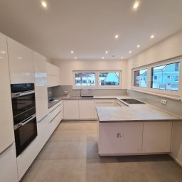 BauTech - Wnętrze nowoczesnej, jasnej kuchni z białymi, lakierowanymi szafkami, szarym blatem i płytkami podłogowymi. Widoczne okna z widokiem na zabudowę jednorodzinną i oświetlenie punktowe w suficie.