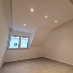 BauTech - Puste, jasne pomieszczenie z oknem, szarą podłogą z płytek, białymi ścianami i sufitem z wbudowanymi punktowymi lampami. Widoczny fragment grzejnika pod oknem.